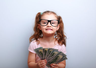 Fun emotional small kid girl in glasses holding and showing dollars. Happy winner on blue background with empty copy space