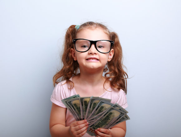 Fun emotional small kid girl in glasses holding and showing dollars. Happy winner on blue background with empty copy space